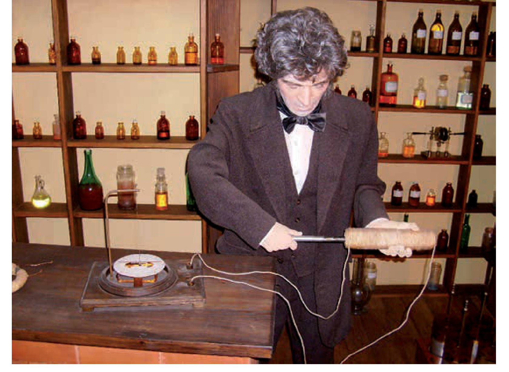 Figura 3: recriação museológica da experiência de Faraday, com o cientista a segurar uma barra magnetizada junto a uma bobina ligada a um galvanómetro, num laboratório com prateleiras de frascos antigos